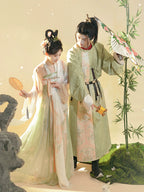 Two individuals in traditional attire standing outdoors with a bamboo plant and stone sculpture in the background.